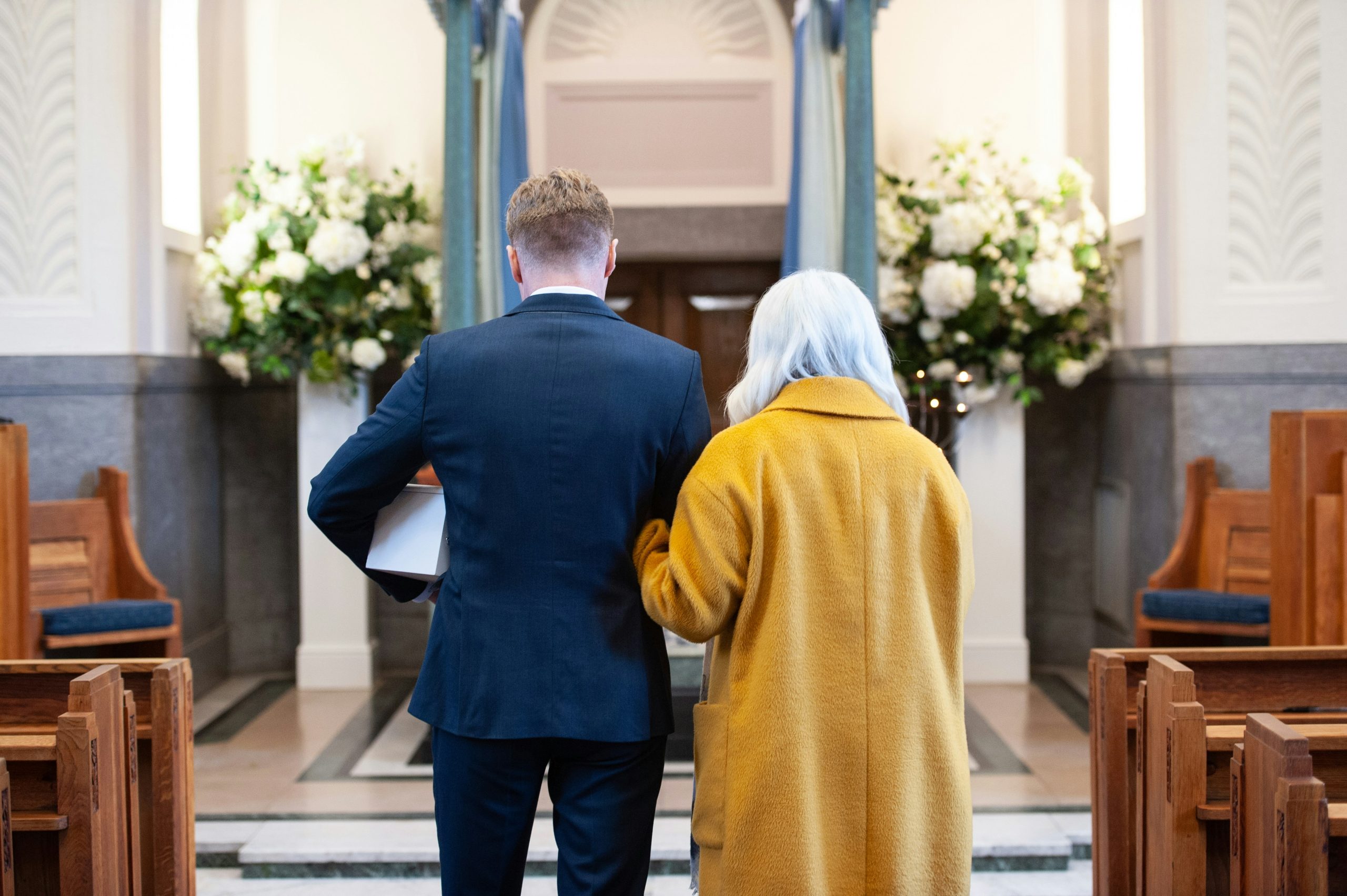 Son and Mom, arms linked, standing, facing toward an alter in a church with pews on either side | Absolute Assignment of Life Insurance and Funeral Funding | Family Funeral Funding - Helping Families with After Death Expenses | Bridging the gap between families, funeral homes and life insurance companies.