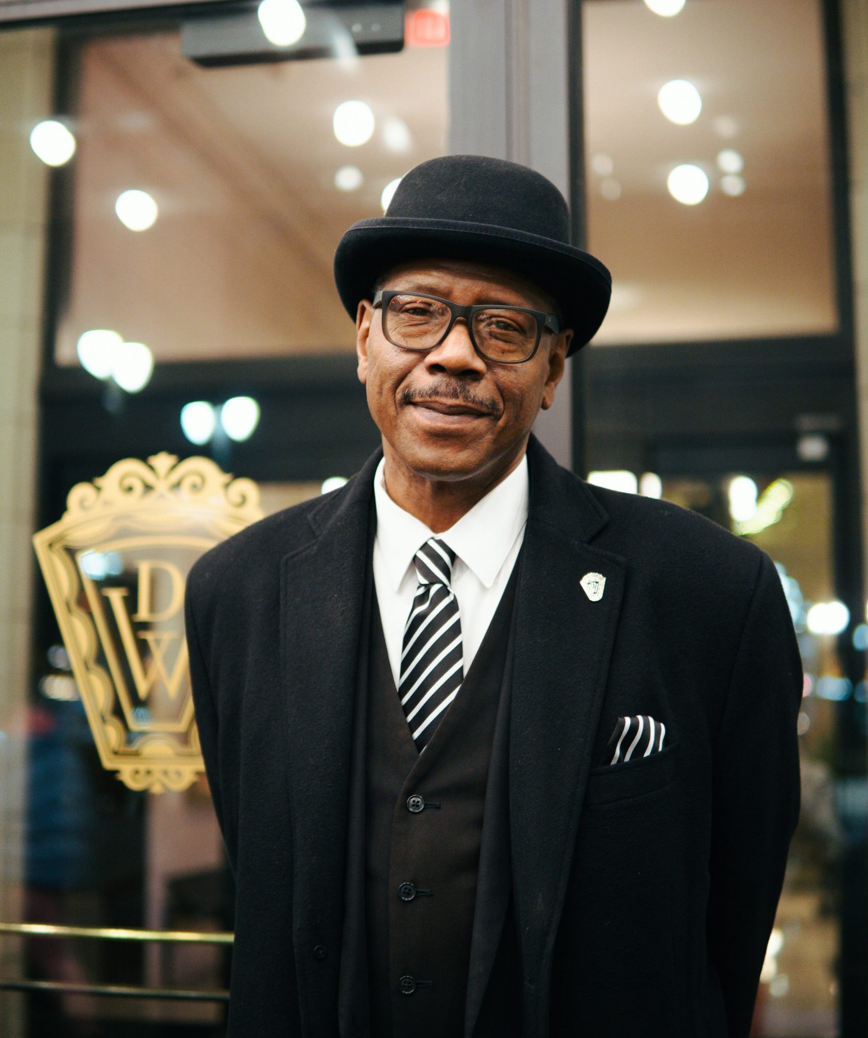 African American funeral director dressed sharply in black with a tie and hat, facing the camera in front of a glass window with a funeral home logo on it | Insurance Assignments: What Funeral Homes Need to Know | Family Funeral Funding - Helping Families with After Death Expenses | Bridging the gap between families, funeral homes and life insurance companies.